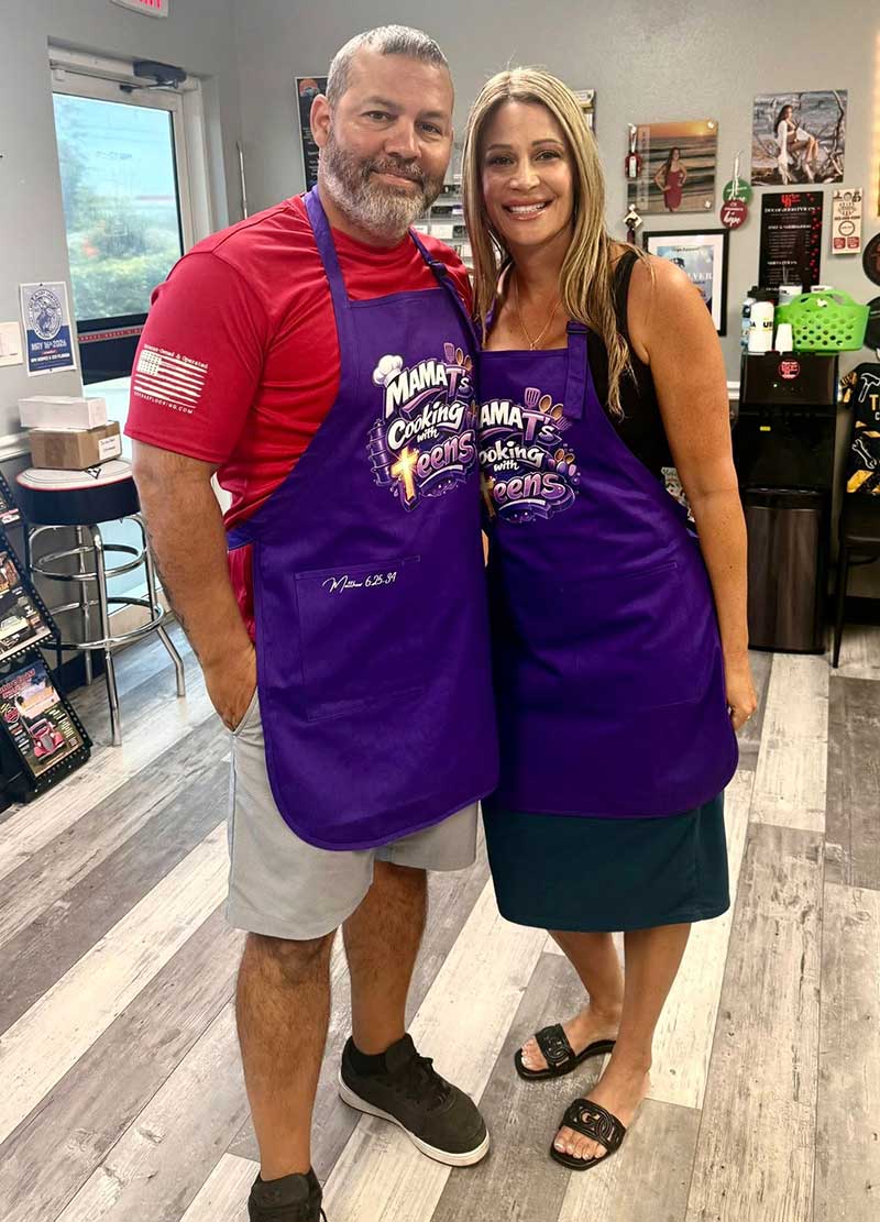 A man and a woman smiling and standing close together indoors, both wearing purple aprons that say "Mama’s Cooking Teams." The man is in a red shirt and gray shorts; the woman is in a dark skirt and light top.
