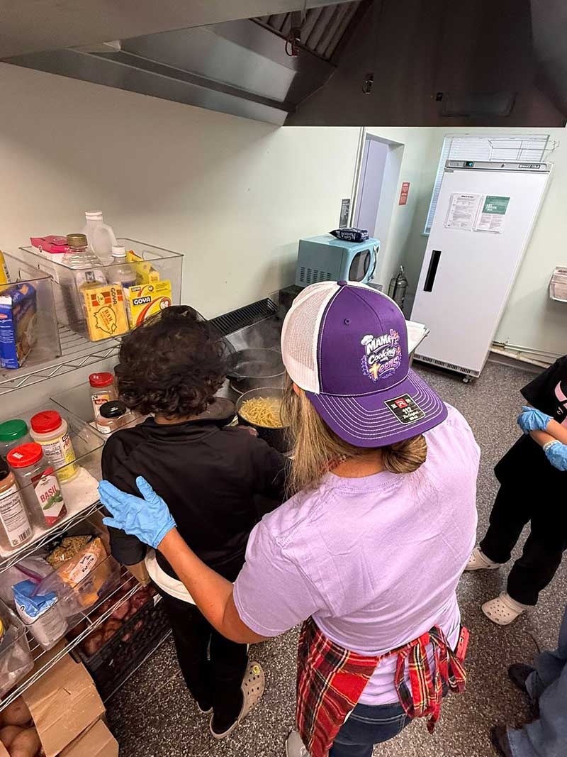 mama-t-cooking-teen Two people stand in a kitchen cooking pasta on a stove. One person, wearing a purple hat and gloves, gently touches the shoulder of another person in black. Shelves with spices and food items are visible nearby.