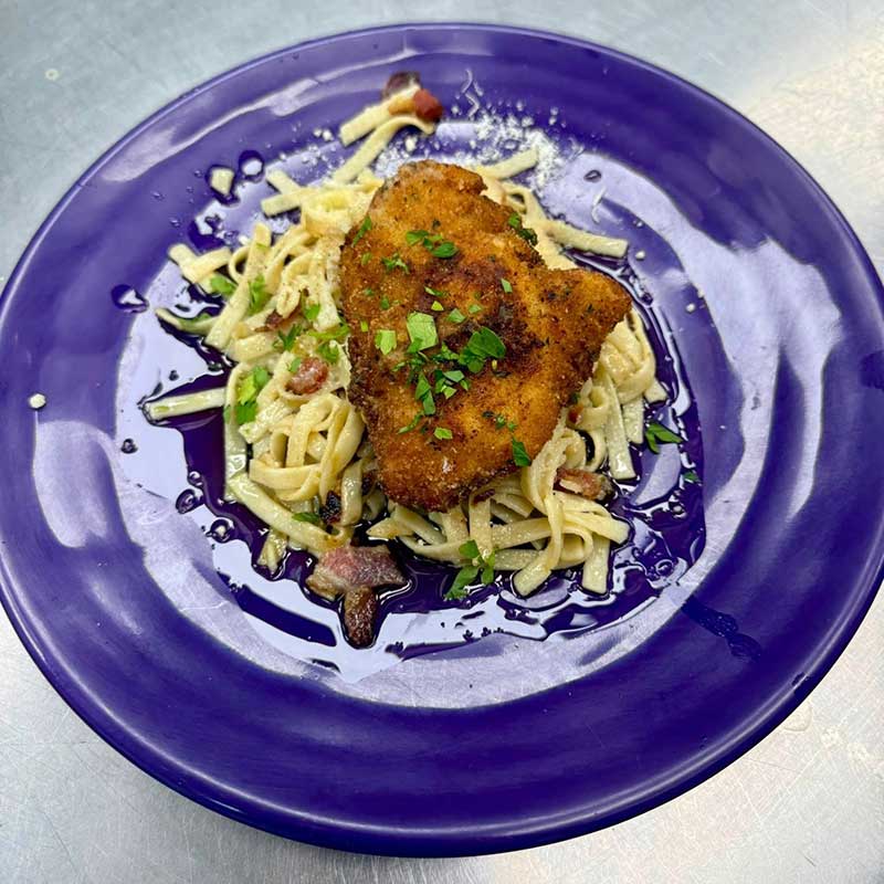 A breaded, fried chicken cutlet sits atop creamy pasta with bits of bacon, garnished with chopped parsley, on a vibrant blue plate.