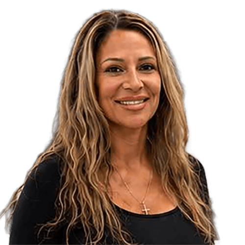 A woman with long wavy light brown hair, wearing a black top and a cross necklace, smiles at the camera against a plain light background.